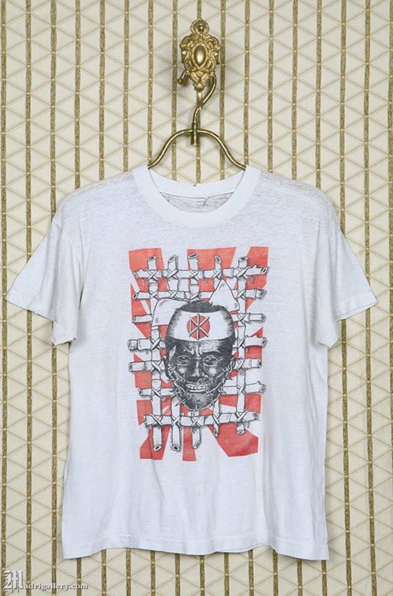 May include: White t-shirt with a black and white graphic print of a person's face with a red circle and a cross on their forehead. The person's face is surrounded by a grid of black lines and the background is red.