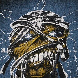 Pu&ograve; includere: Una maglietta blu con una grafica di un teschio giallo e nero con bende bianche e catene. Il teschio ha occhi blu luminosi e un'espressione minacciosa. Il testo "THE CHURCH OF SATAN.COM" &egrave; stampato nell'angolo in basso a destra della grafica.