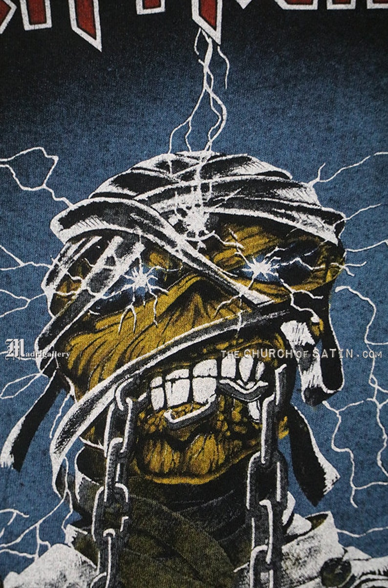 Pu&ograve; includere: Una maglietta blu con una grafica di un teschio giallo e nero con bende bianche e catene. Il teschio ha occhi blu luminosi e un'espressione minacciosa. Il testo "THE CHURCH OF SATAN.COM" &egrave; stampato nell'angolo in basso a destra della grafica.