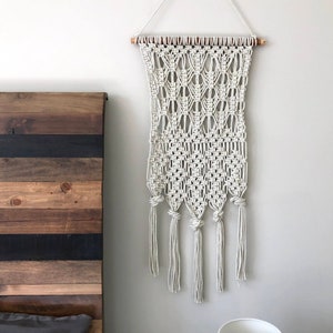May include: A white macrame wall hanging with a geometric pattern and tassels. The hanging is attached to a wooden dowel rod and is hung on a white wall.