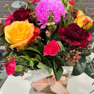May include: A bouquet of flowers in a brown box with a white and orange striped ribbon. The flowers include a yellow rose, pink carnations, red carnations, and a purple chrysanthemum.