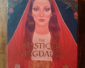 May include: The Mystic of Magdalene Oracle of Love deck box features a woman with long brown hair wearing a red robe and holding a golden chalice. The box has a dark blue background with gold accents and the text "The Mystic of Magdalene An Oracle of Love".