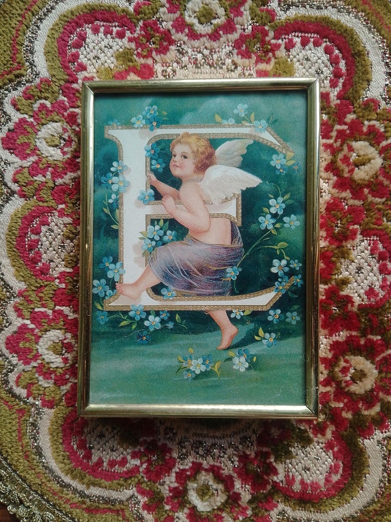 May include: A framed vintage print of a cherub angel with wings, holding onto a large white letter E, surrounded by blue forget-me-not flowers on a green background.