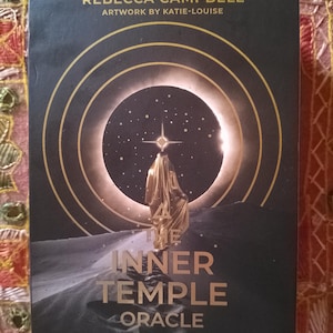 Può includere: Scatola di carte oracolo nera con scritte e design dorati. Il testo sulla scatola recita "REBECCA CAMPBELL ARTWORK BY KATIE-LOUISE" e "THE INNER TEMPLE ORACLE". L'immagine sulla scatola presenta una figura in abiti dorati.