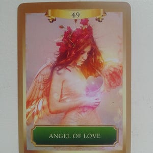 May include: Angel of Love card with a golden border and a green banner with the text "Angel of Love". The card features a woman with red hair and wings holding a pink heart.