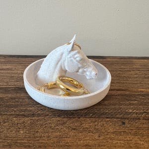 May include: White ceramic horse head ring dish with gold hoop earrings. The dish has a speckled finish.