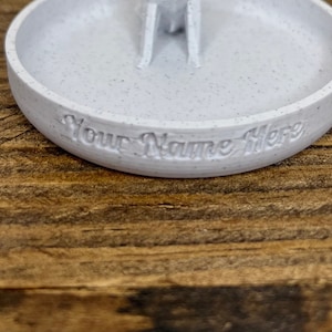 May include: White ceramic ring dish with a small raised center and the text "Your Name Here" inscribed on the edge.