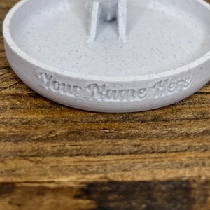 May include: A white ceramic ring dish with a speckled finish. The dish has a small indentation in the center and the words "Your Name Here" are inscribed on the bottom.