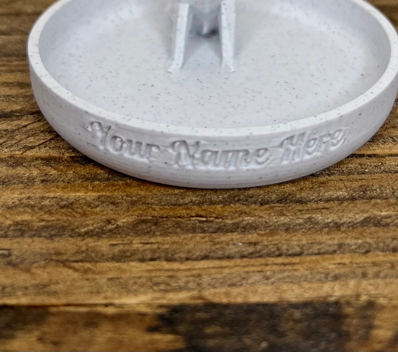 May include: A white ceramic ring dish with a speckled finish. The dish has a small indentation in the center and the words "Your Name Here" are inscribed on the bottom.