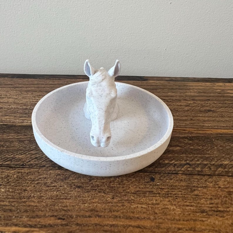 May include: White ceramic ring dish with a horse head design. The dish is speckled with small white flecks.
