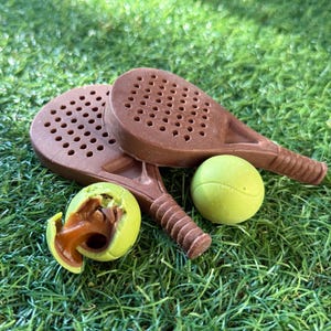 May include: Two brown padel rackets with a green tennis ball and a broken green ball revealing a caramel filling, on green grass. The rackets have a perforated head and a textured handle. The rackets and balls are likely chocolate.