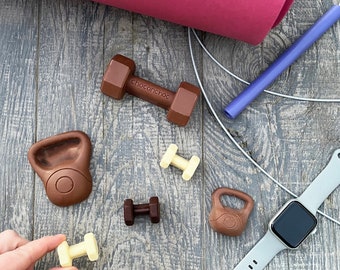 May include: A collection of chocolate gym equipment, including dumbbells, a kettlebell, and a yoga mat. The chocolate is brown and has the words "chocochoc" on one of the dumbbells.