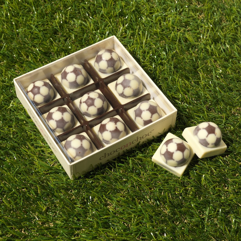 Puede incluir: Una caja de nueve bolas de f&uacute;tbol de chocolate blanco y marr&oacute;n en una caja blanca con el texto "chocochoc" en ella. La caja est&aacute; sentada sobre una superficie verde cubierta de hierba.