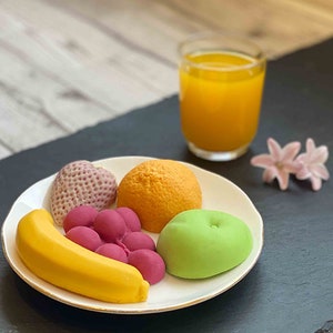 Op de afbeelding: Een wit bord met een verscheidenheid aan kleurrijke fruitvormige zeepjes. De zeepjes zijn gevormd als een banaan, sinaasappel, appel, druiven en een hartvormige aardbei. Het bord staat op een zwart oppervlak naast een glas sinaasappelsap.