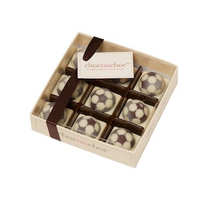 Puede incluir: Una caja de bombones de chocolate blanco con forma de bal&oacute;n de f&uacute;tbol. La caja es marr&oacute;n y blanca con el texto "chococonchoc&trade; Lovingly handmade in Great Britain" en la parte delantera.