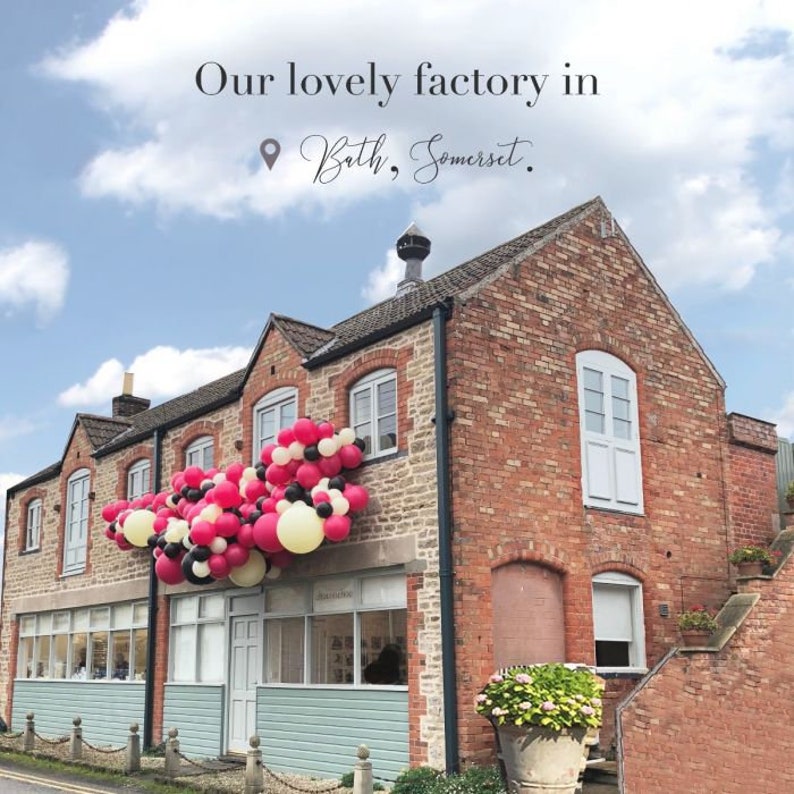 Op de afbeelding: Een bakstenen gebouw met een grote ballonboog gemaakt van roze, zwarte en witte ballonnen. Het gebouw heeft een bord met de tekst "Onze mooie fabriek in Bath, Somerset."