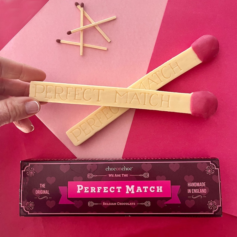 Puede incluir: Dos cerillas de chocolate blanco con puntas rojas, una con el texto "PERFECT MATCH" y la otra con el texto "PERFECT" sobre un fondo rosa y blanco. Las cerillas est&aacute;n al lado de una caja con el texto "choconchoc WE ARE THE PERFECT MATCH BELGIAN CHOCOLATE" y "THE ORIGINAL" y "HANDMADE IN ENGLAND".