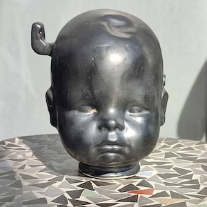 May include: A black ceramic sculpture of a baby's head with a small horn on the side. The sculpture is sitting on a table with a mosaic tile top.