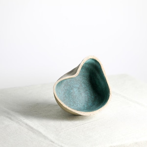 May include: A small, heart-shaped ceramic bowl with a white exterior and a teal blue interior. The bowl is sitting on a white linen tablecloth.