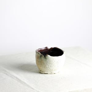 May include: A small, white ceramic bowl with a rough, textured surface. The bowl has a dark brown interior and a slightly uneven rim.