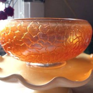 May include: An orange glass bowl with a textured surface. The bowl is on a white pedestal with a scalloped edge.