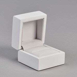 May include: A white ring box with a hinged lid, open to reveal a white velvet interior with a ring pillow.