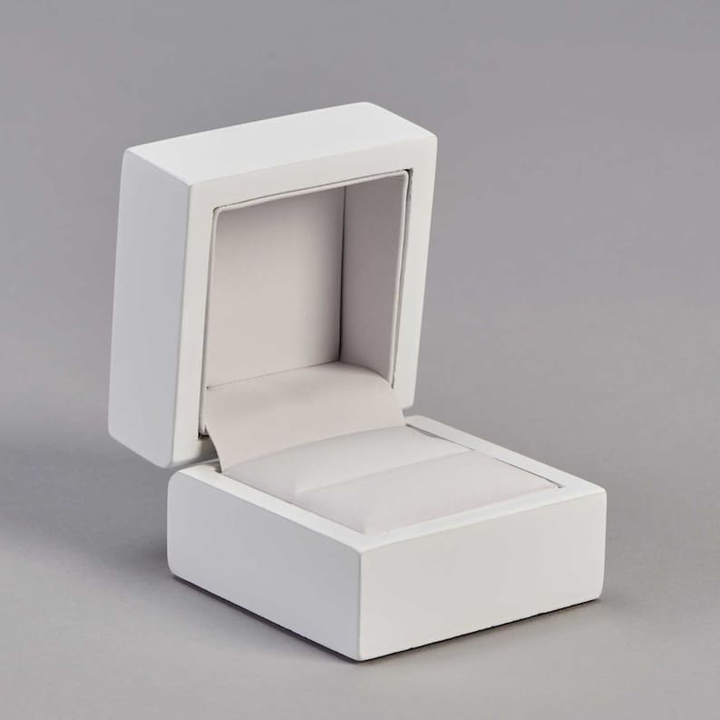 May include: A white ring box with a hinged lid, open to reveal a white velvet interior with a ring pillow.
