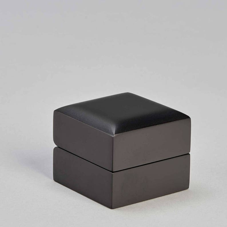 May include: A black ring box with a hinged lid. The box is made of a smooth, matte material and has a simple, elegant design.