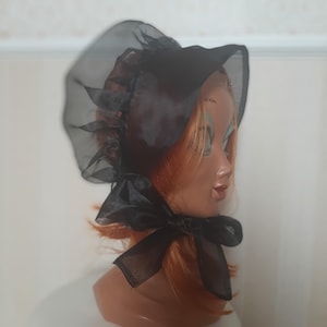 May include: A black bonnet made of sheer, translucent fabric featuring a ruffled edge and a wide ribbon tie that secures under the chin. The headwear is displayed on a mannequin head with reddish-brown hair, showing the semi-transparent texture.