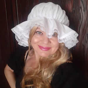 Op de afbeelding: Een witte kantmuts met een geplooide ruche rond de kroon. De muts wordt gedragen door een persoon met blond haar en is schuin naar de zijkant gekanteld.
