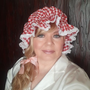 Op de afbeelding: Een vrouw draagt een wit blouse met een rood-wit geruite muts. De muts heeft witte kantversiering. Ze heeft blond haar en draagt een roze lint in haar haar.