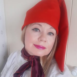 Può includere: Un cappello da elfo rosso brillante con una lunga punta. Il cappello è indossato con una sciarpa bordeaux e una camicetta bianca. Una piccola campana d'argento pende dalla punta del cappello. L'immagine è un primo piano di una persona che indossa il cappello e la sciarpa.