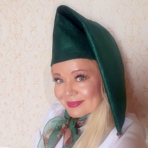 May include: A dark green, pointed hat with a long, draped side. The hat is made of a soft, velvety material. The hat is worn by a person with blonde hair and a white shirt.