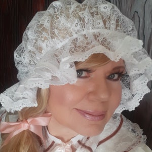 Op de afbeelding: Een witte kantmuts met een gerimpelde rand. De muts wordt gedragen door een persoon met blond haar en make-up.