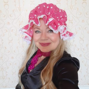 Op de afbeelding: Een vrouw draagt een roze en wit gebloemde muts met witte kant. Ze draagt ook een zwart satijnen blouse en een roze satijnen sjaal.