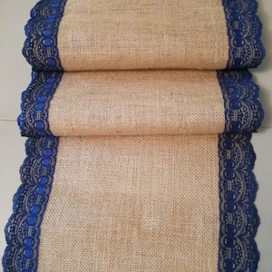 Op de afbeelding: Een opgevouwen tafelloper van jute met een donkerblauwe kanten rand langs de randen. De loper heeft een natuurlijke bruine kleur en het kant voegt een decoratief tintje toe. De tafelloper is waarschijnlijk voor de woondecoratie.