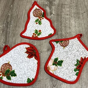 Könnte beinhalten: Drei weiße Topflappen mit Weihnachtsmotiven und rotem Rand. Die Topflappen sind in Form eines Herzens, eines Baumes und eines Hauses. Jeder Topflappen hat ein anderes Design mit einem Weihnachtsornament, Stechpalme und Weihnachtsstern.