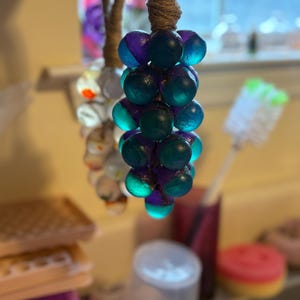 May include: A cluster of decorative orbs in shades of teal and purple, suspended from a natural twine cord. The orbs have a glossy finish and are arranged to resemble grapes. The background is blurred, suggesting a kitchen setting.