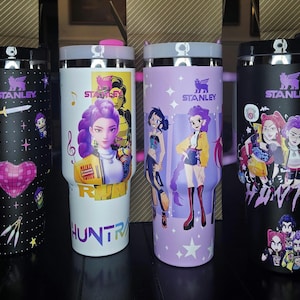May include: Four Stanley tumblers with black, white, and purple designs. Each tumbler features a black lid and a graphic design. The designs include illustrations of characters, a heart, a sword, and the word "HUNTRA". The Stanley logo is visible on each tumbler.