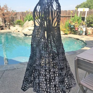 Puede incluir: Un chaleco de crochet negro con un patrón de tejido suelto y abierto. El chaleco se muestra en un maniquí de metal negro sobre un fondo de piscina y patio.