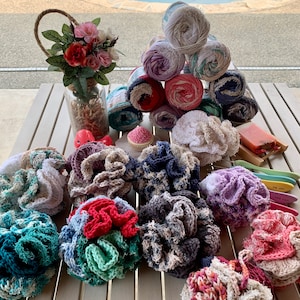 May include: A collection of colorful crocheted bath poufs in various colors, including blue, green, red, and white. The poufs are arranged on a wooden table with a vase of flowers and yarn balls in the background.