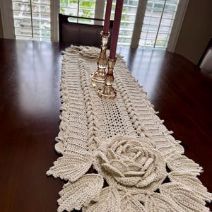 May include: A beige crocheted table runner with a large rose and leaf design. Two gold candle holders with burgundy candles sit on the runner. The runner is on a dark wood table.