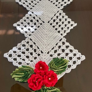 May include: White crocheted table runner with a scalloped edge and three red crocheted flowers with green leaves.