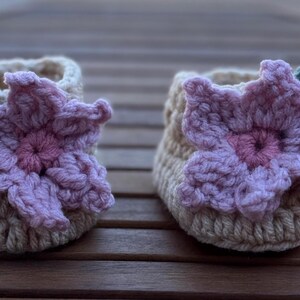 May include: A pair of handmade crochet baby booties. Each bootie is beige with a large, light purple flower and a green leaf accent. The booties are designed with a textured stitch and are suitable for a newborn.