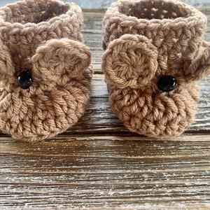 May include: A pair of brown crocheted baby booties with black button eyes. The booties have a simple design and are perfect for a newborn.
