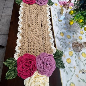 May include: A rectangular crochet table runner in a tan cable-knit pattern with a white scalloped border. The runner is decorated with large, three-dimensional crocheted flowers in shades of cream, magenta, and mauve, accented by green leaves.