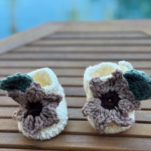 May include: A pair of handmade crochet baby booties. The booties are off-white with a brown and cream-colored flower and green leaf embellishments. The booties are displayed on a wooden surface.