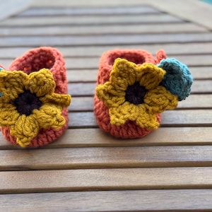 May include: A pair of handmade crochet baby booties in a warm orange hue. Each bootie features a large, yellow sunflower with a dark brown center. A small teal accent is attached to one of the booties. The booties are placed on a wooden surface.