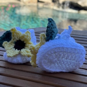 May include: Two white crochet baby booties with sunflower and leaf accents. One bootie features a yellow and black sunflower, while the other has a green leaf. The booties are handmade and sit on a wooden surface, perfect for a baby shower gift.
