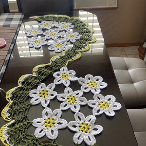 May include: A crocheted table runner with a green and yellow border and white flowers with yellow centers.